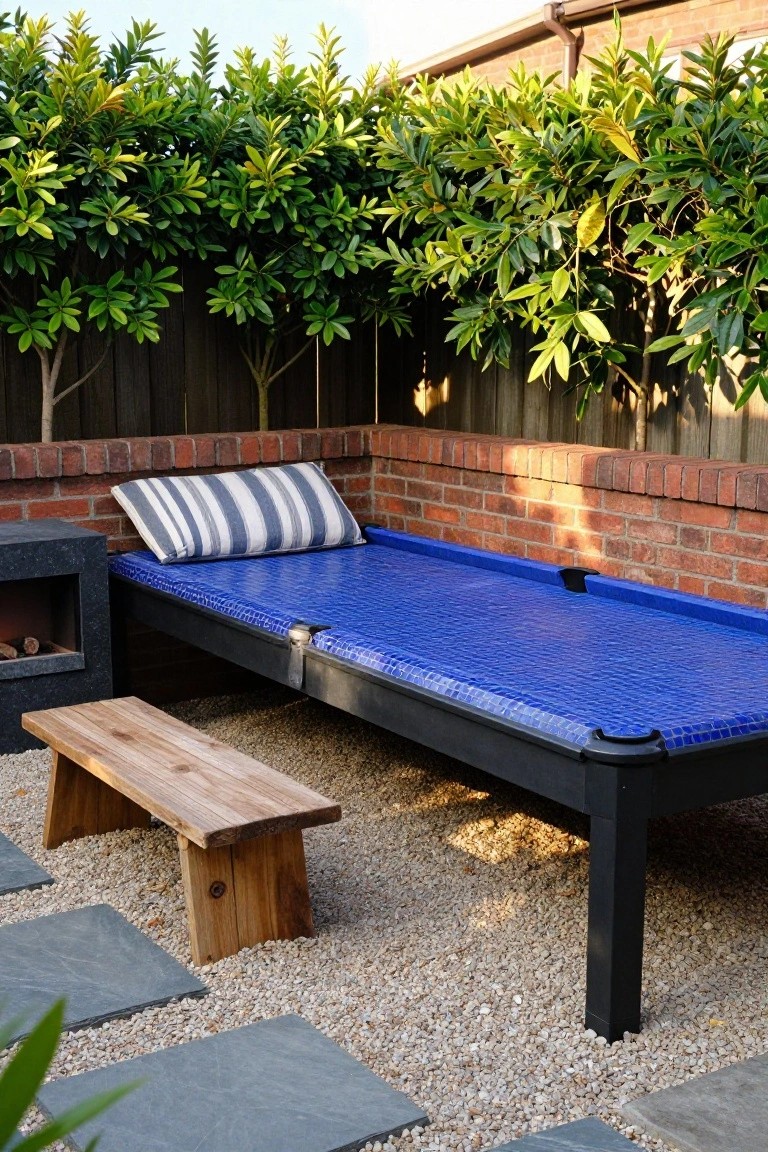 Blue mosaic-tiled pool platform used as an outdoor daybed with a striped cushion, next to a modern stone fire pit and wooden bench in a gravel backyard area with brick wall, greenery hedge, and stepping stones.