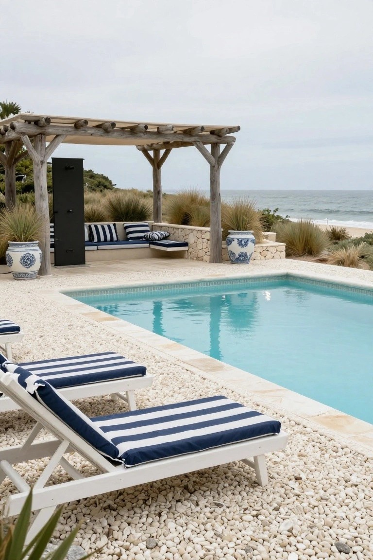 20 Luxury Pool Exterior Ideas That Boost Curb Appeal Turquoise rectangular pool edged in light stone and surrounded by white pebbles, with two white loungers having navy blue striped cushions nearby, a rustic wooden pergola sheltering a built-in bench with matching cushions, blue and white ceramic pots, stone walls, and ocean dunes in the background.