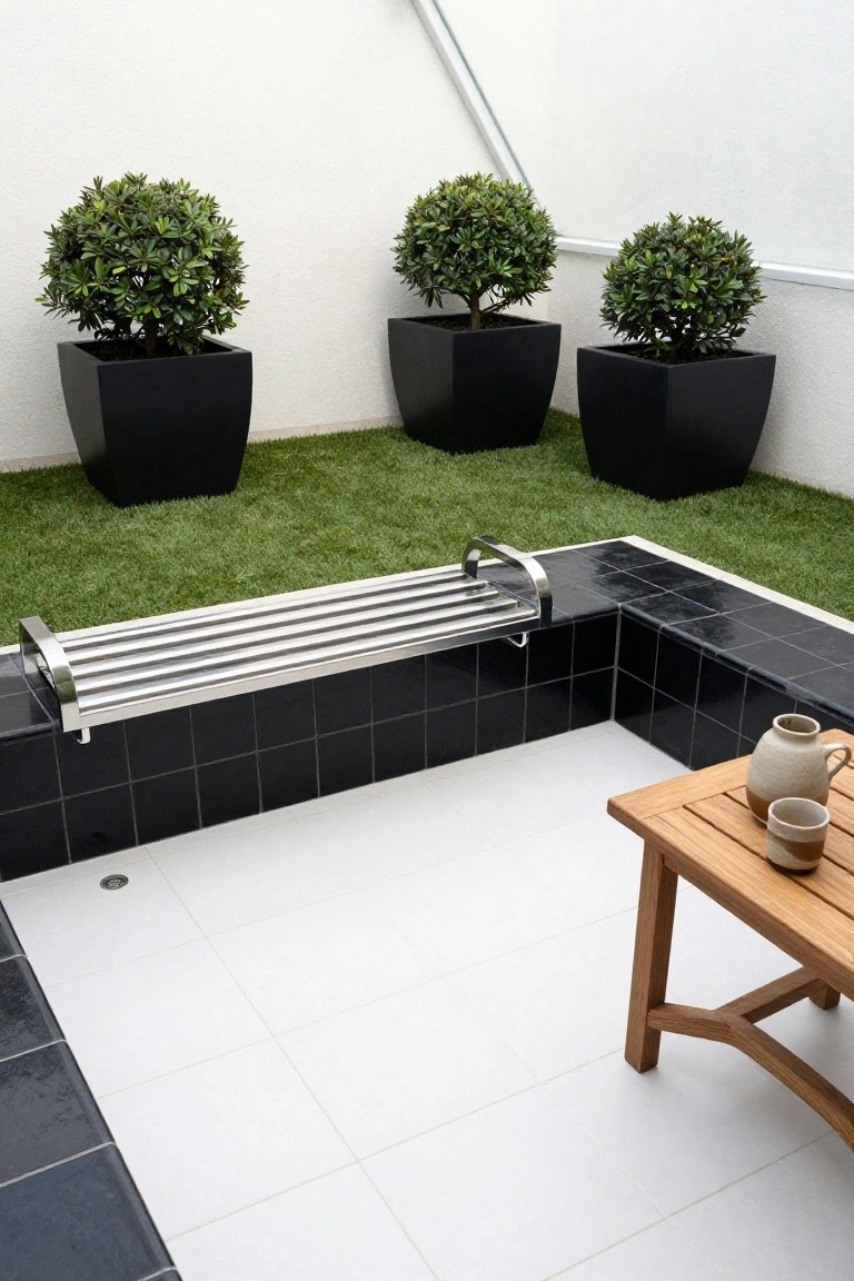 Small plunge pool with black tiled edges and integrated stainless steel slatted bench along one side, surrounded by artificial turf, three black pots with boxwood plants, and a wooden table holding ceramic pots and cups on white tiled patio.
