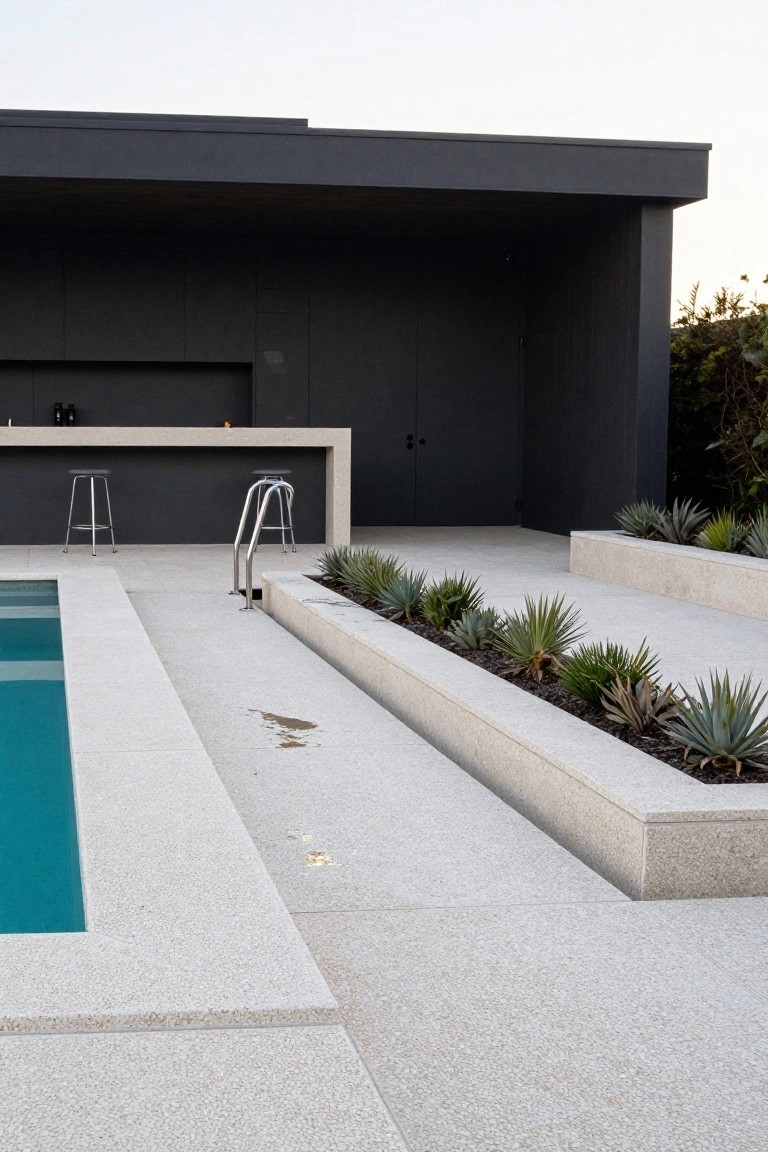 Turquoise pool with stainless steel ladder, bordered by white aggregate decking and linear raised concrete planters filled with agave succulents, adjacent to an outdoor concrete bar with stools against a dark paneled wall.