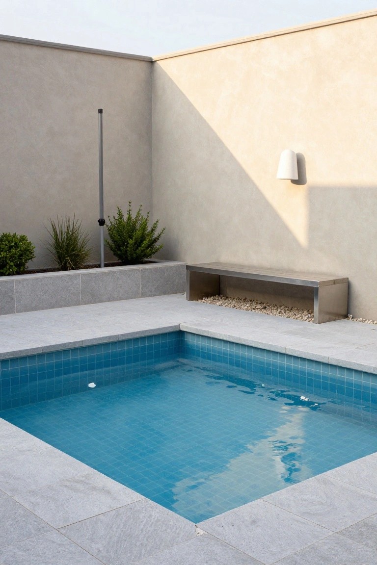 Small square plunge pool with blue mosaic tiles and clear water, surrounded by light gray stone pavers, a long stainless steel bench positioned alongside, low green plants in a raised bed, beige rendered walls, and a slim metal pole nearby.