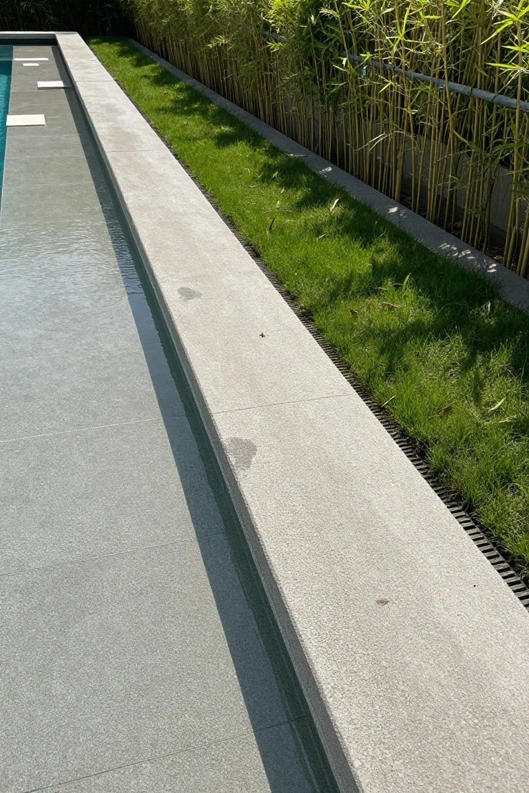 Close-up of a rectangular infinity-edge pool with gray concrete coping, a narrow strip of green grass adjacent to the deck, and dense tall bamboo plants screened by a metal rail behind a concrete retaining wall.