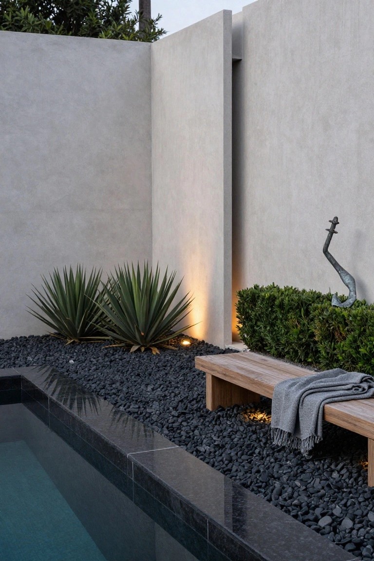 Gray concrete walls beside a narrow pool edged in black granite tiles, with black gravel mulch bed holding two agave plants, a boxwood hedge, metal sculpture, wooden bench draped in gray blanket, and ground lights.