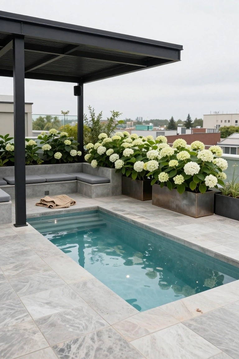 20 Practical Pool Shade Canopy Ideas For Hot Summer Days Rooftop terrace with small turquoise plunge pool, gray marble-look tiled decking, L-shaped concrete bench seating with gray cushions, large white hydrangea plants in rectangular metal planters, black metal canopy structure overhead, and urban buildings in the background.