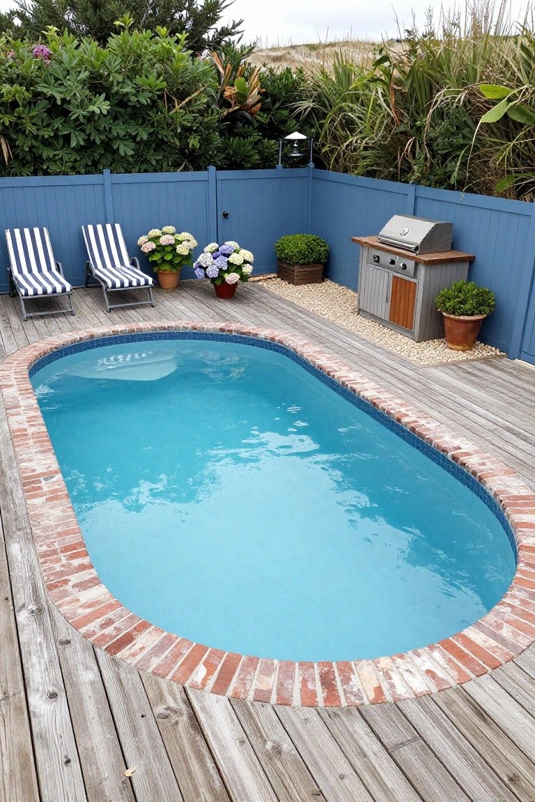 Oval semi-inground pool with brick coping on a wooden deck surrounded by blue privacy fence, lounge chairs, potted hydrangeas, boxwood shrubs, and a built-in grill with gravel edging.