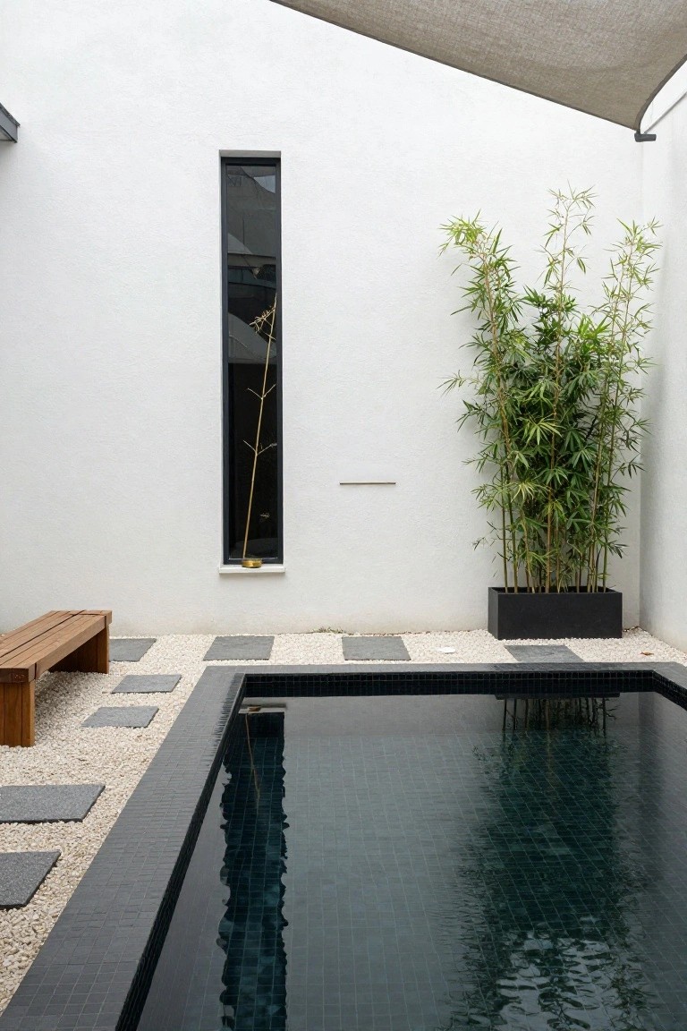 Modern courtyard with a rectangular dark mosaic-tiled pool, white walls featuring a narrow black-framed window with a golden vase inside, tall bamboo in a black planter, wooden bench, pebble groundcover, and dark stone pavers.