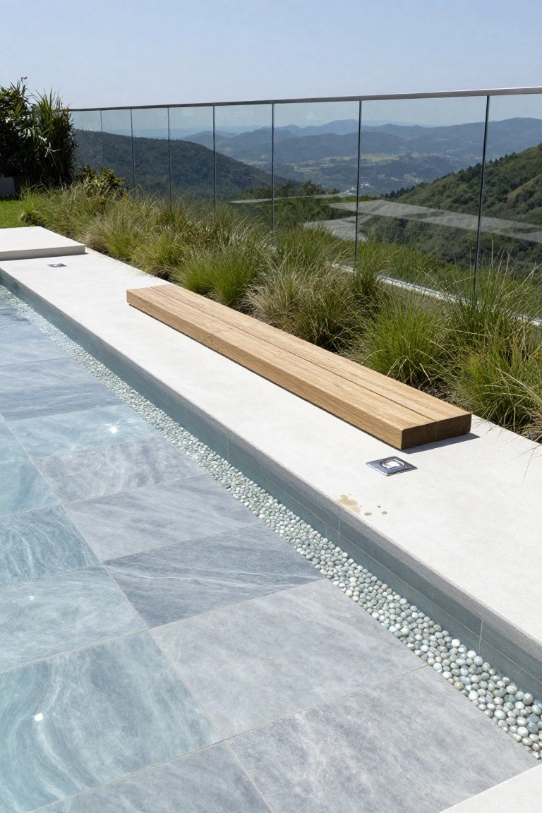 Infinity-edge swimming pool with blue-gray veined marble tiles, white pebble edging, wooden bench nearby, ornamental grasses, and distant mountain view beyond glass railing.