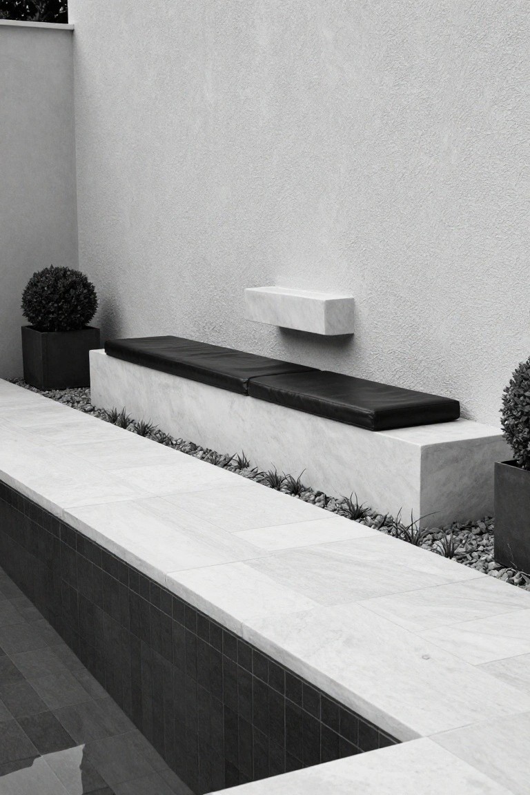 Black and white photo of a half-inground pool with black tiled interior, white marble coping, and a built-in white marble bench topped with black leather cushions along the edge, with boxwood plants in black pots and gravel ground cover nearby.