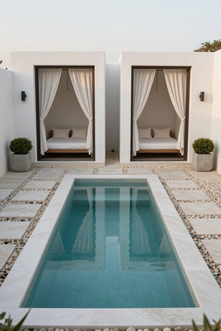 Narrow turquoise plunge pool centered between two white open-air cabanas containing platform beds with white linens and sheer curtains, surrounded by light stone pavers and pebbles with potted plants nearby.