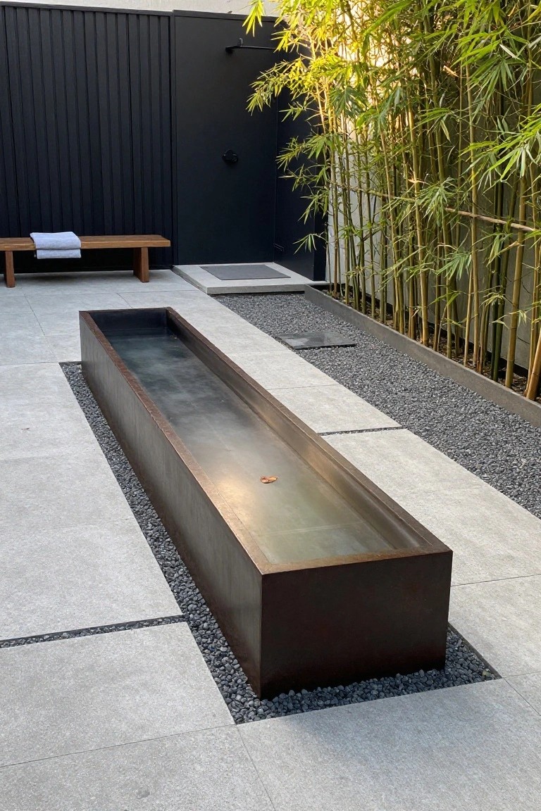 Long rectangular corten steel pool in an outdoor patio surrounded by light gray pavers, black gravel borders, tall bamboo plants, a wooden bench with towels, and a black outdoor shower enclosure.