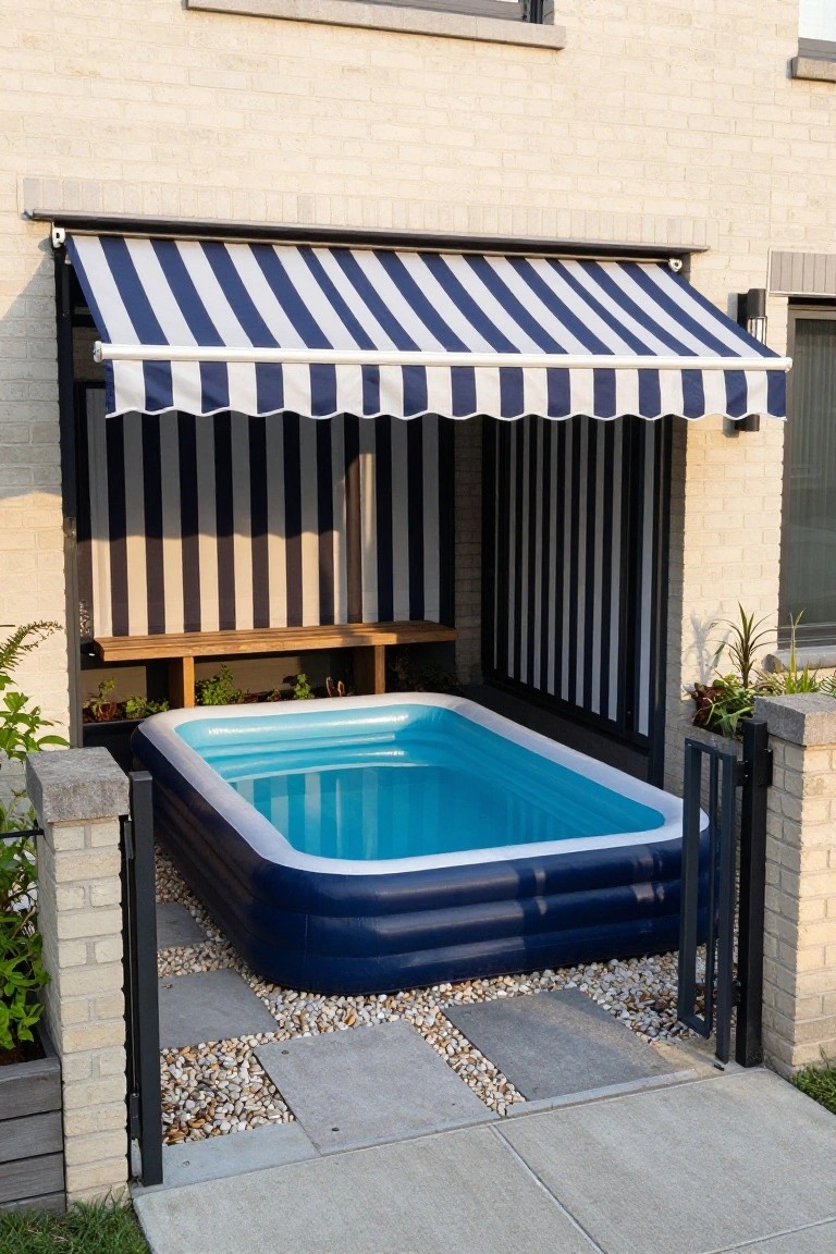 Small blue inflatable kiddie pool under blue and white striped retractable awning attached to brick house exterior, with wooden bench, plants, gravel ground, and metal gate nearby.