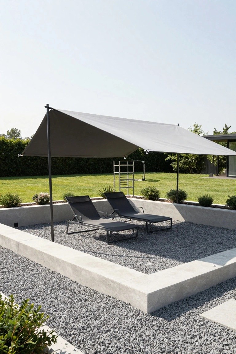 Gray cantilevered shade sail covering two black lounge chairs on a gravel patio surrounded by concrete retaining walls, plants, and a grassy lawn.