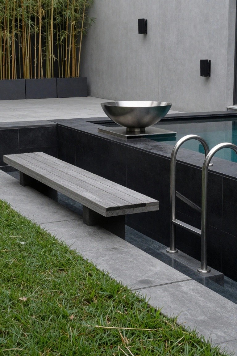 21 Inspiring Pool Deck Makeover Ideas You’ll Want to Try This Summer A modern pool deck features a long wooden slat bench along the black tiled pool edge next to a stainless steel ladder, with a large metallic bowl on a pedestal nearby, bamboo plants in black planters, gray concrete paving, and green grass.