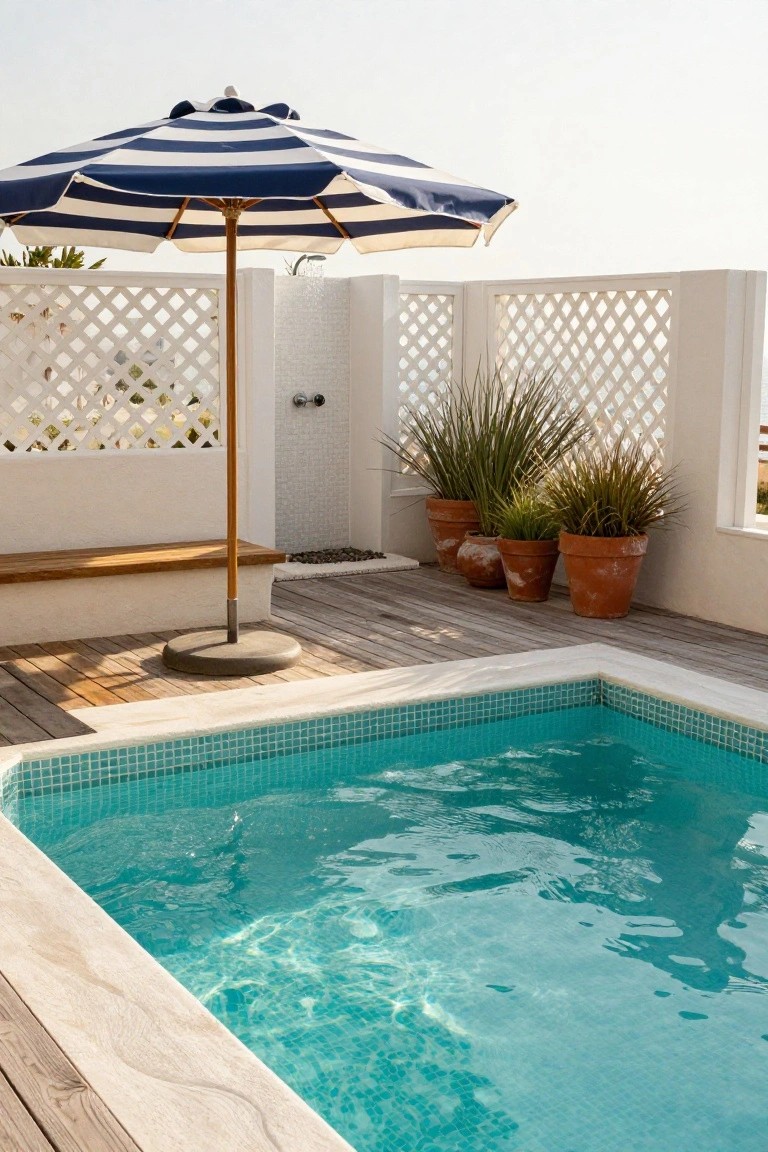 Turquoise mosaic-tiled inground pool with white tiled edges on a wooden deck, adjacent to a striped umbrella, outdoor shower, lattice screens, and potted plants.
