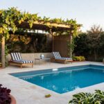 Wooden pergola draped in green climbing vines with bamboo screen walls shades two striped chaise lounges and a side table beside a pool, with potted plants in terracotta pots along the edge.