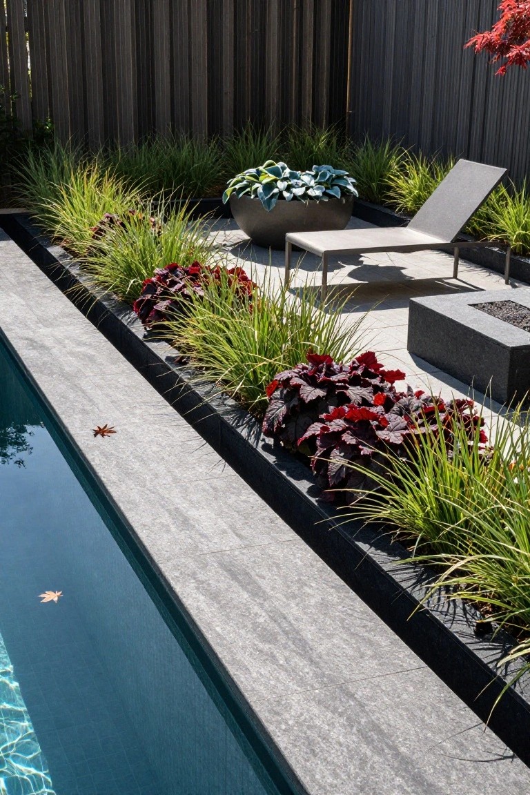 Sleek pool edge with black metal planters filled with green ornamental grasses and burgundy coleus foliage, a lounge chair nearby, a square gas fire pit, and wooden fence backdrop.