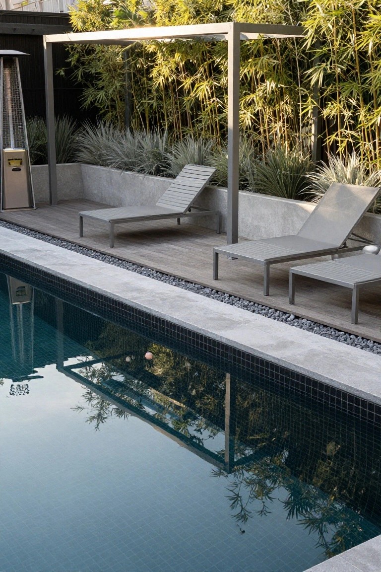 Inground pool with turquoise water, black tile edges, adjacent wood deck with three gray metal loungers, pebble strip border, concrete raised planters filled with grasses and bamboo screen on metal frame.