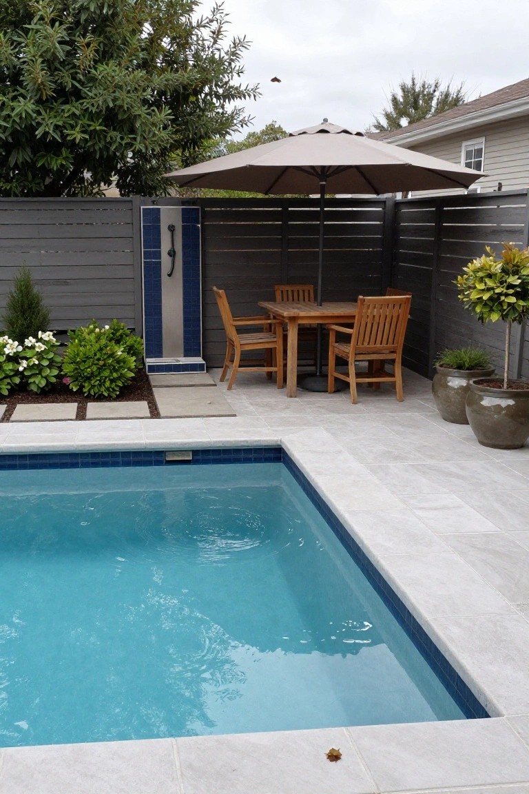 Small rectangular inground pool with blue tiled edges surrounded by light gray pavers, teak wood dining table and four chairs under a beige umbrella next to an outdoor shower on a slatted gray fence, with potted plants and shrubs nearby.