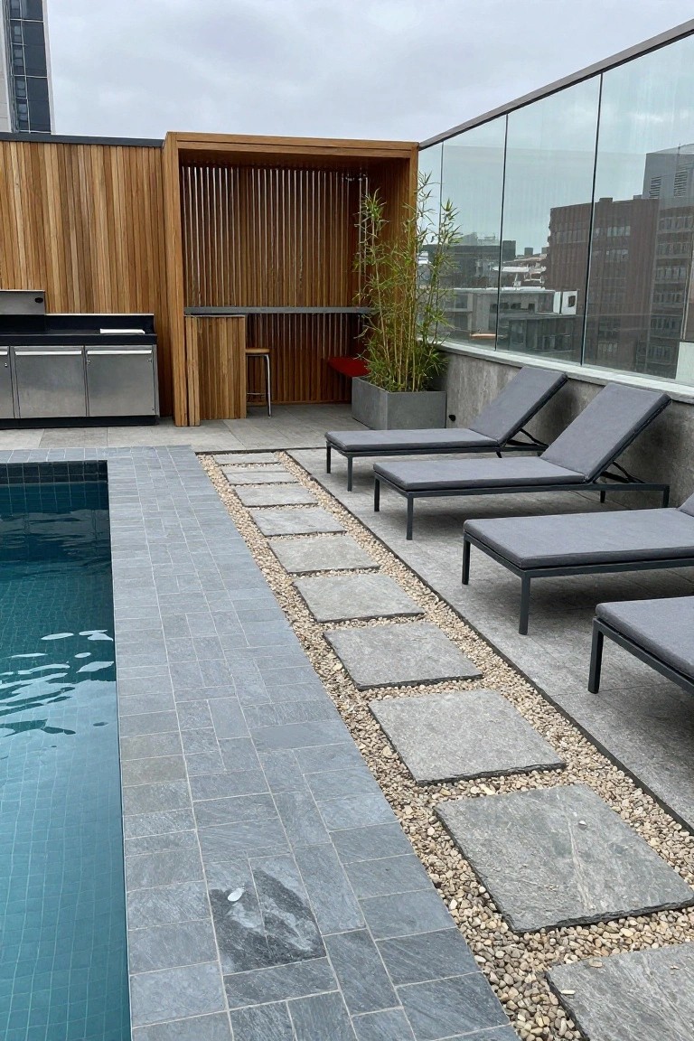 Rooftop pool area with dark blue mosaic pool tiles, dark gray slate surround tiles, pebble-edged stepping stones leading to gray lounge chairs, stainless steel BBQ, wooden bar with bamboo plants, and glass railings.