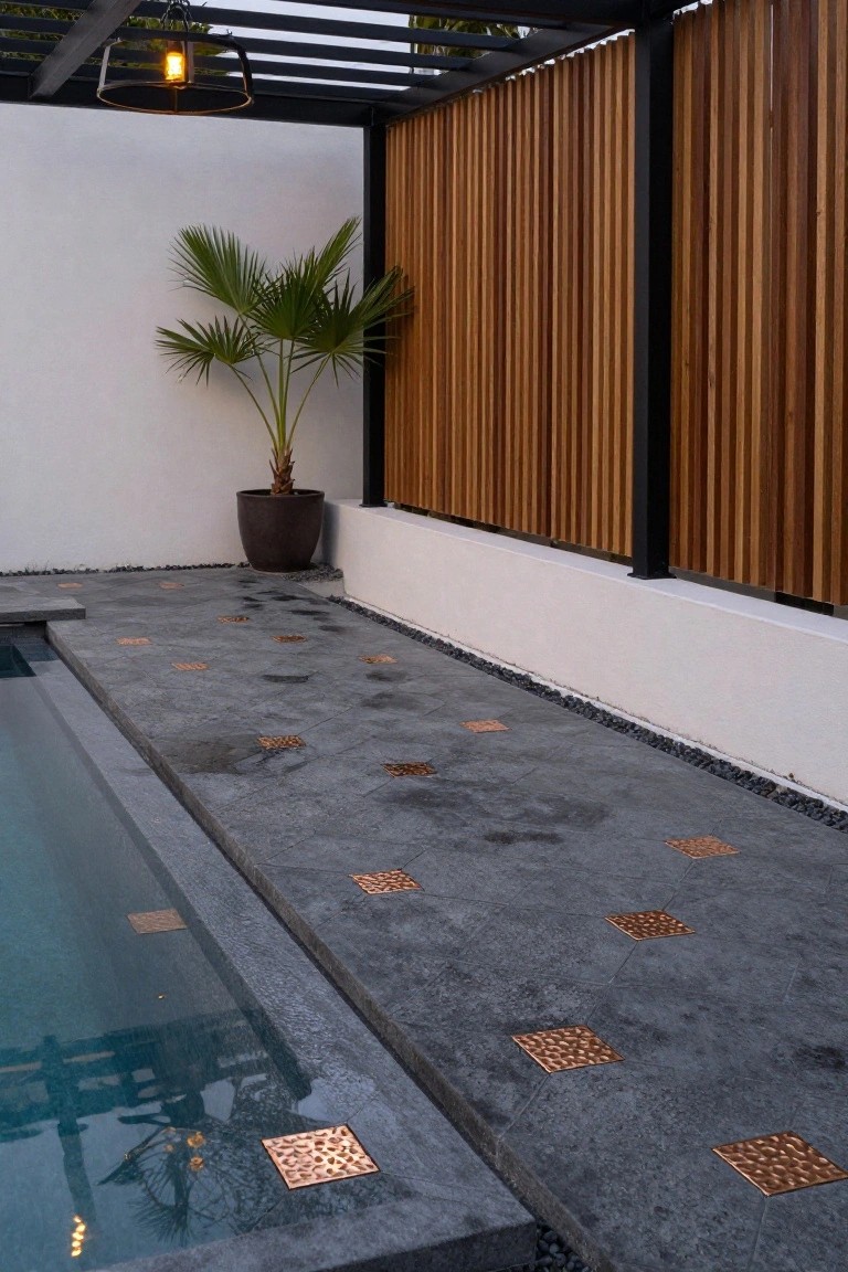 Pool deck made of dark gray stone tiles with hammered copper square inserts embedded along the edge next to a rectangular pool, featuring black metal pergola with hanging light, vertical wood slat fencing, white wall, potted palm tree, and gravel border.