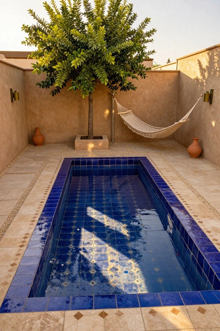 Small rectangular pool lined with blue patterned tiles, surrounded by light beige deck tiles featuring pebble-filled borders and inlays, with a hammock under a tree in a walled outdoor space and terracotta pots nearby.