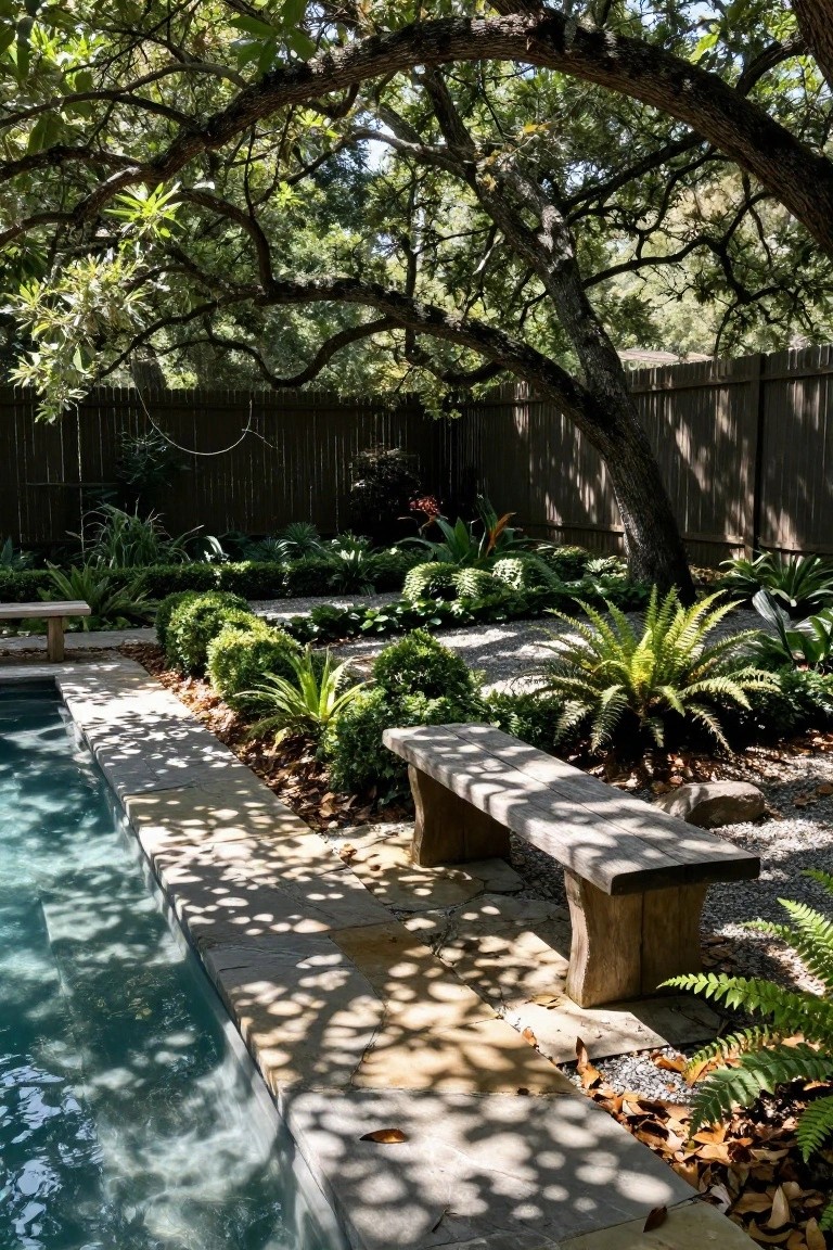 Backyard pool with light blue water along a stone paver edge, surrounded by gravel paths, low boxwood hedges, ferns, and other tropical plants, a wooden bench nearby, and overhanging branches from large trees casting shadows.