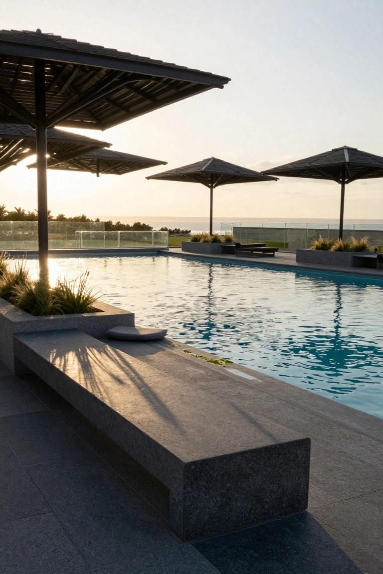 Rooftop pool at sunset with multiple large thatched umbrellas clustered nearby, stone benches and potted grasses along the edge, lounge chairs, and glass railing overlooking ocean and grass.