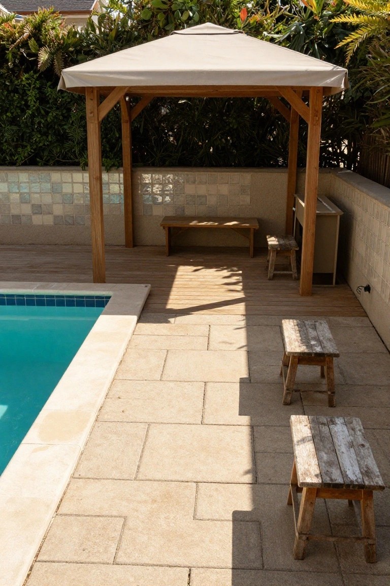 Wooden pergola with beige canopy over a bench, adjacent to a small wooden cabinet and rustic stools, next to a blue semi-inground pool with beige stone coping and light stone pavers, backed by greenery and a tiled wall.