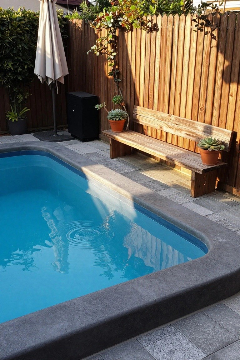 Semi-inground pool with clear blue water and black edging on a stone tiled deck, next to a rustic wooden bench with potted plants, white umbrella, and wooden fence in the background.