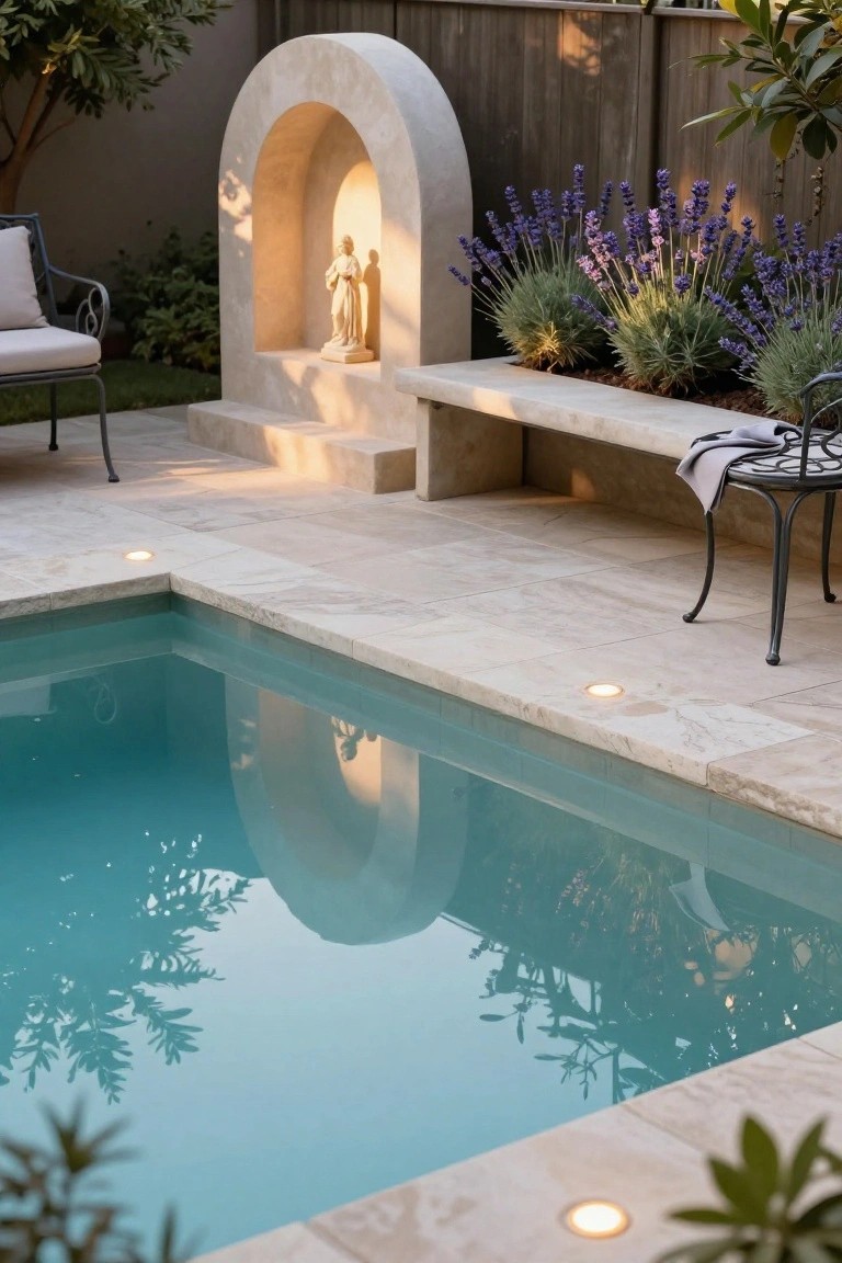 Small rectangular pool with travertine tile surround and underwater edge lights glowing yellow, arched stone niche holding a statue nearby, lavender plants in raised beds, wooden fence, and two metal chairs on the patio.