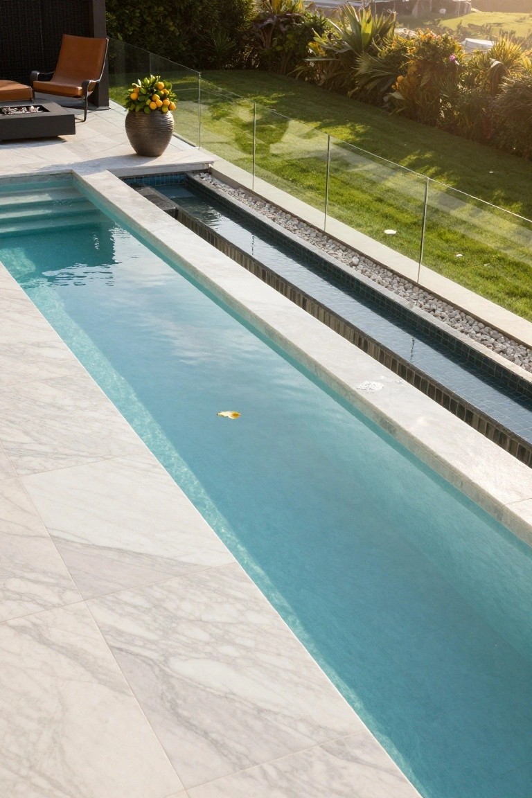 Narrow lap pool with turquoise water and white marble decking surrounded by glass railings, pebble spillover channel, potted orange tree, leather chair, fire pit, and lush green landscaping.