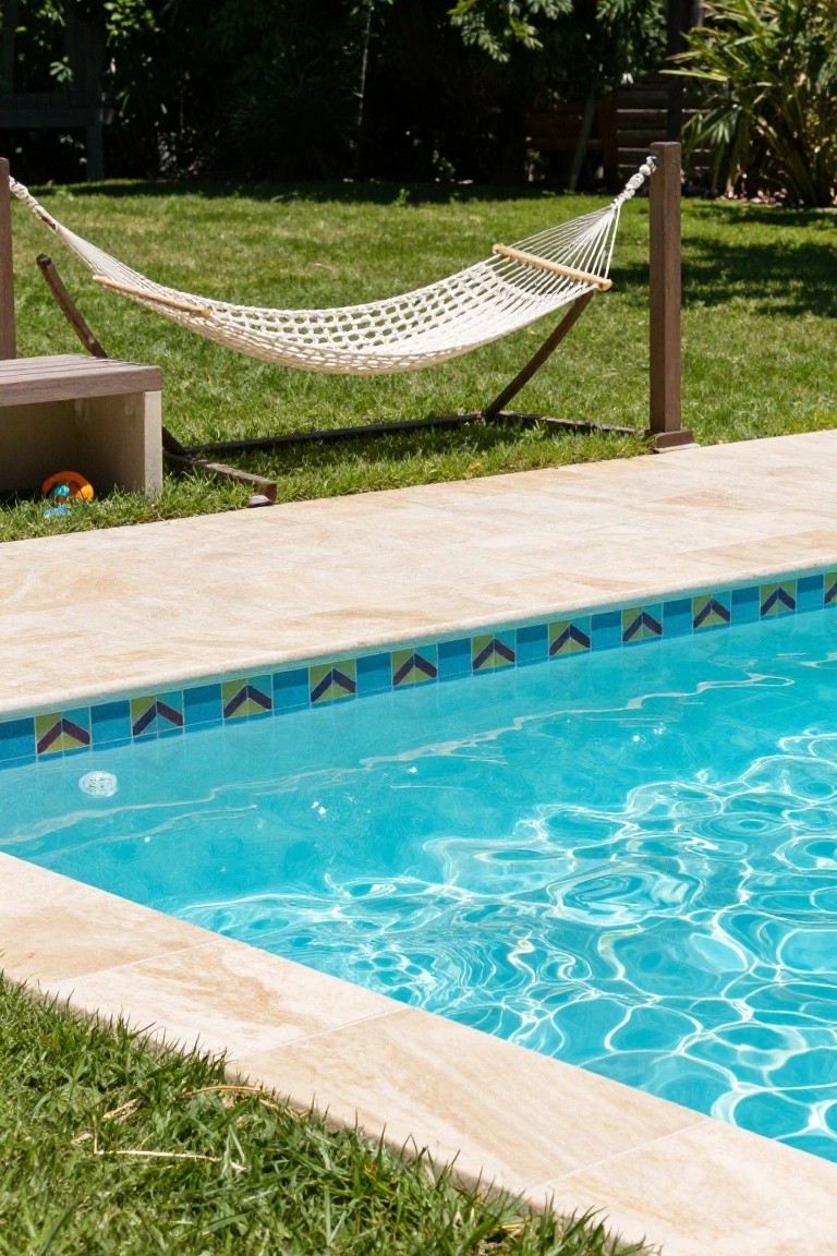 Backyard swimming pool with beige travertine decking and a border of blue, green, and black chevron mosaic tiles along the edge, white hammock stand and grass nearby.