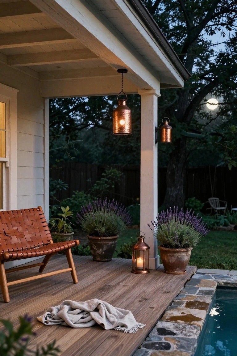 Wooden porch deck with woven leather chair, potted lavender plants, copper hanging lanterns and freestanding lantern, overlooking stone-edged pool at dusk with trees, fence, and moon in background.