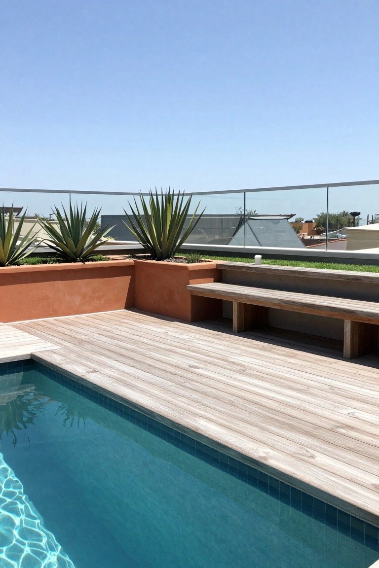 Small semi-inground pool on a light wooden deck adjacent to a raised orange planter wall with agave plants and an integrated wooden bench seat, plus glass railing and distant rooftops under blue sky.