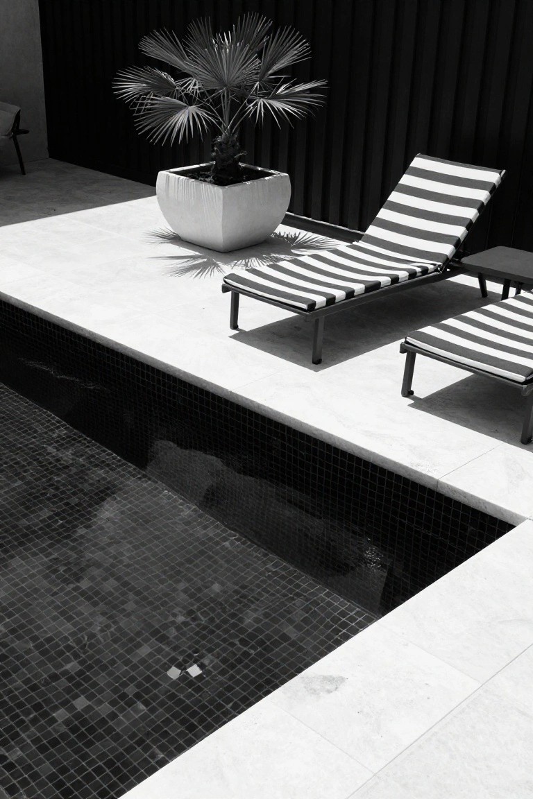 Black and white photo of a pool area with black mosaic-tiled pool, white stone deck, two black-and-white striped loungers, potted palm plant, and tall black fence in the background.