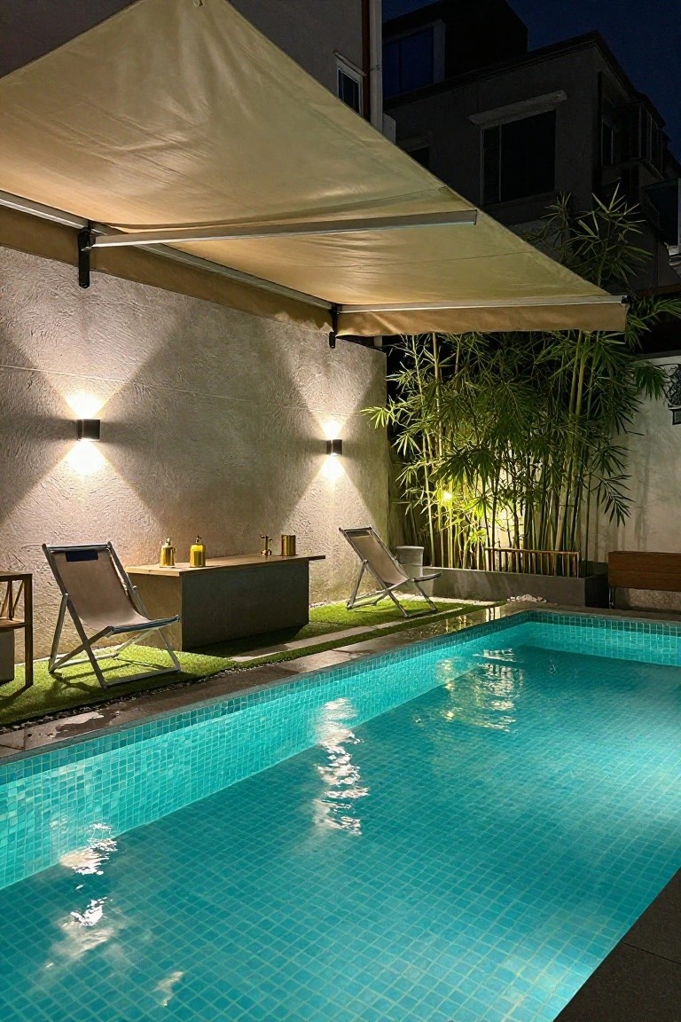 Nighttime backyard pool area with beige retractable awning overhead, two lounge chairs beside a low table with bottles, square wall-mounted lights on textured wall, bamboo plants in corner planter, and glowing turquoise mosaic-tiled pool.