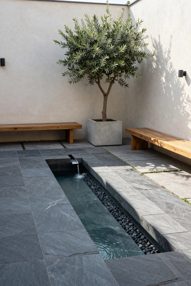 Narrow rectangular raised pool with flowing water over pebbles in a dark slate-tiled courtyard, flanked by wooden benches and a potted olive tree against white walls.