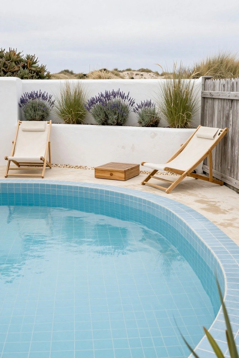 Curved turquoise-tiled raised swimming pool with two adjacent wooden lounge chairs with white cushions on a light deck, lavender plants and grasses along white walls and wooden fence, dunes visible in background.