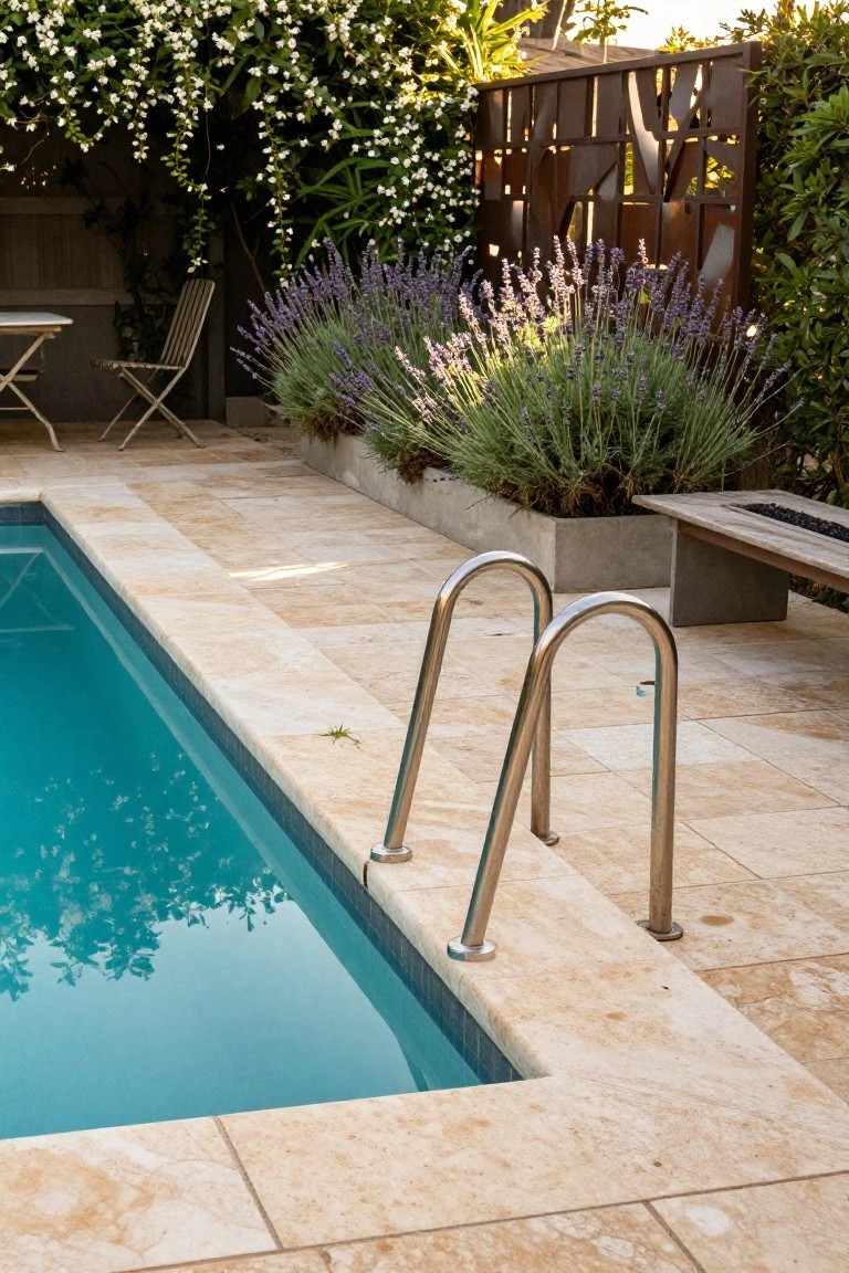 Narrow rectangular turquoise pool with curved stainless steel ladder, surrounded by beige travertine tiles, adjacent wooden fire pit bench, concrete planter with grasses, lavender shrubs, and climbing white flowers on a backyard patio.