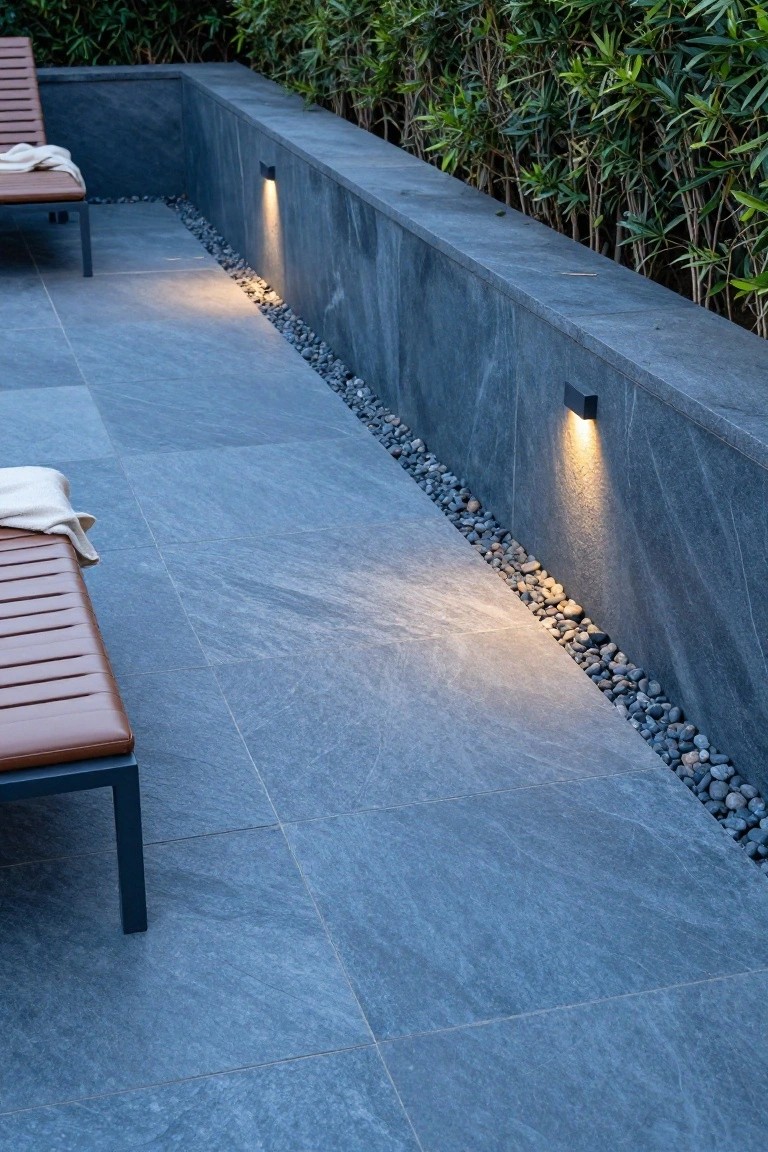 Outdoor terrace with large blue-gray slate-look tiles, tan wood-frame loungers with beige cushions and white towels, pebble gravel border along a dark slate wall with black wall-mounted lights, and green bamboo hedge backdrop.