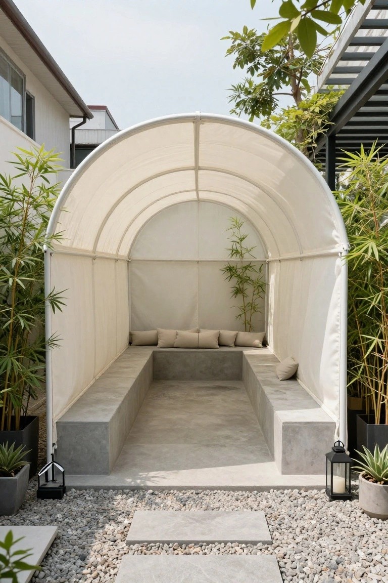 White arched canvas canopy covering a U-shaped concrete bench seating area with beige cushions, surrounded by bamboo plants, pebble ground cover, stone steps, and black lanterns in a garden courtyard.