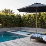Pool deck with two blue-and-white striped lounge chairs under a large beige cantilever umbrella, beside a small rectangular swimming pool edged in concrete, bamboo plants and glass railing in the background.