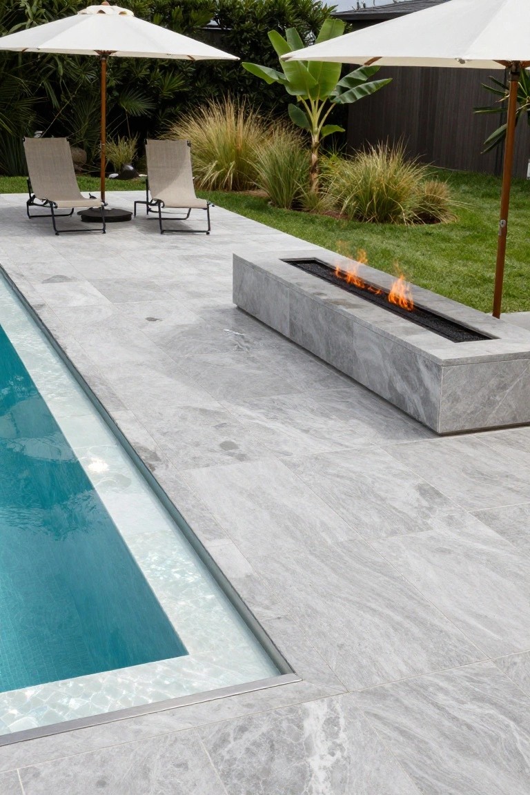 Gray veined stone tiled pool deck with built-in linear gas fire pit along the edge, turquoise pool water, lounge chairs under white umbrellas, tropical plants and grass in the background.