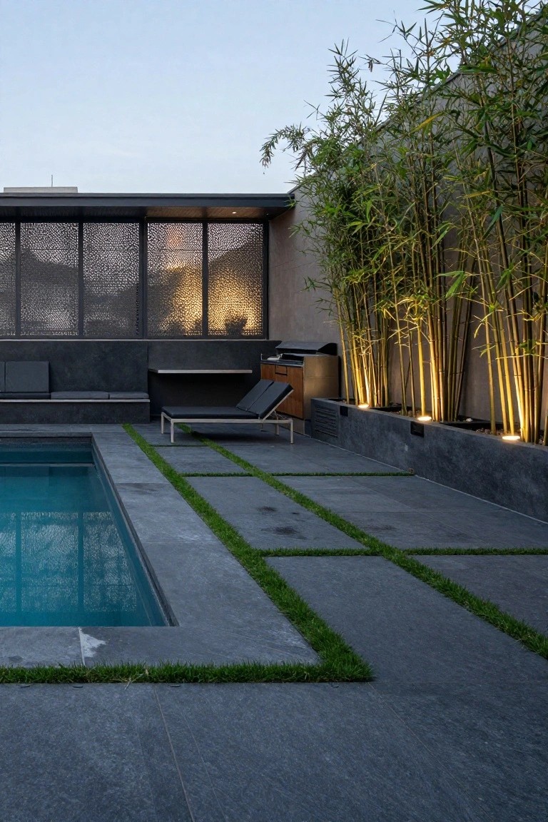 Rooftop pool deck featuring dark gray pavers with linear green grass insets along the pool edge, flanked by tall bamboo plants with uplighting, a lounge chair, built-in seating, and barbecue station.