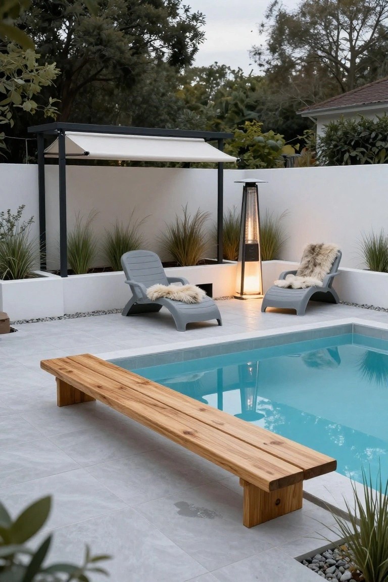 A turquoise pool with a long wooden bench along the edge, flanked by gray loungers with sheepskin throws, tall patio heater, potted grasses, white walls, and pebble accents on a tiled patio.