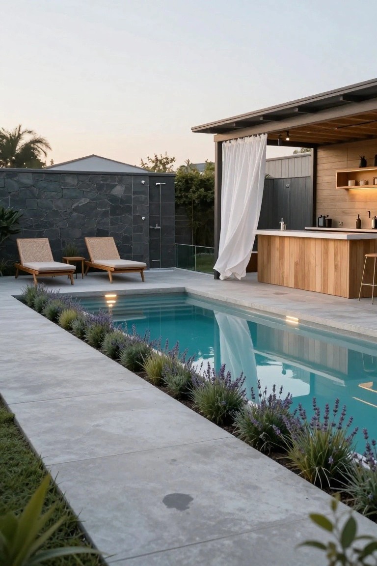 Narrow turquoise inground pool edged with lavender plants and grasses, flanked by concrete paving, wooden loungers, and an adjacent wooden outdoor bar with white curtains and stools in a backyard.