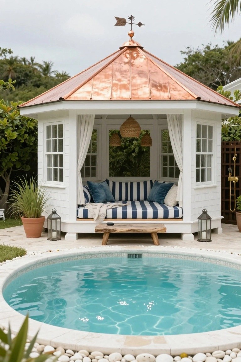 White octagonal gazebo with copper roof, sheer white curtains, rattan hanging lanterns, blue-striped cushions on a wooden daybed bench, potted plants and lanterns nearby, adjacent to a small round turquoise pool edged in stone and pebbles with tropical landscaping.