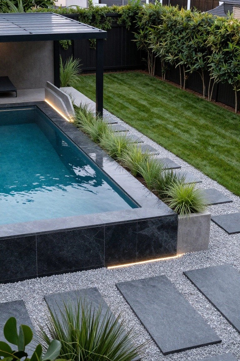 Rectangular lap pool with black marble tiled surround and LED strip lighting under the edge, ornamental grasses planted along one side in concrete planters, dark stone slab pavers set in white pebble path adjacent, green lawn and hedges beyond.