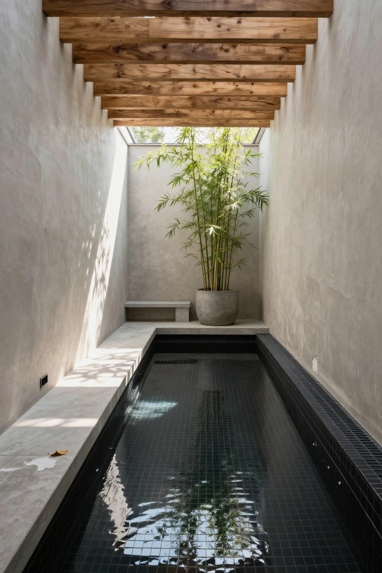 Narrow rectangular indoor pool with black mosaic tiles, concrete benches along one side, a tall potted bamboo plant nearby, light gray plaster walls, wooden ceiling beams, and natural light from a skylight.
