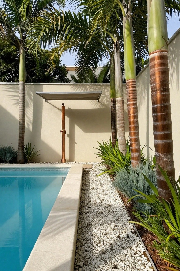 Turquoise pool edged with white pebble mulch bed planted with blue agave, grasses, and palm trees, plus a copper outdoor shower post under a beige wall and awning.